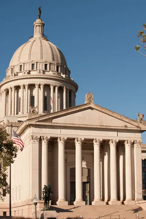 Columned south portico and dome of the Oklahoma State Capitol under a clear blue sky, with the U.S. flag and a bronze statue at the steps