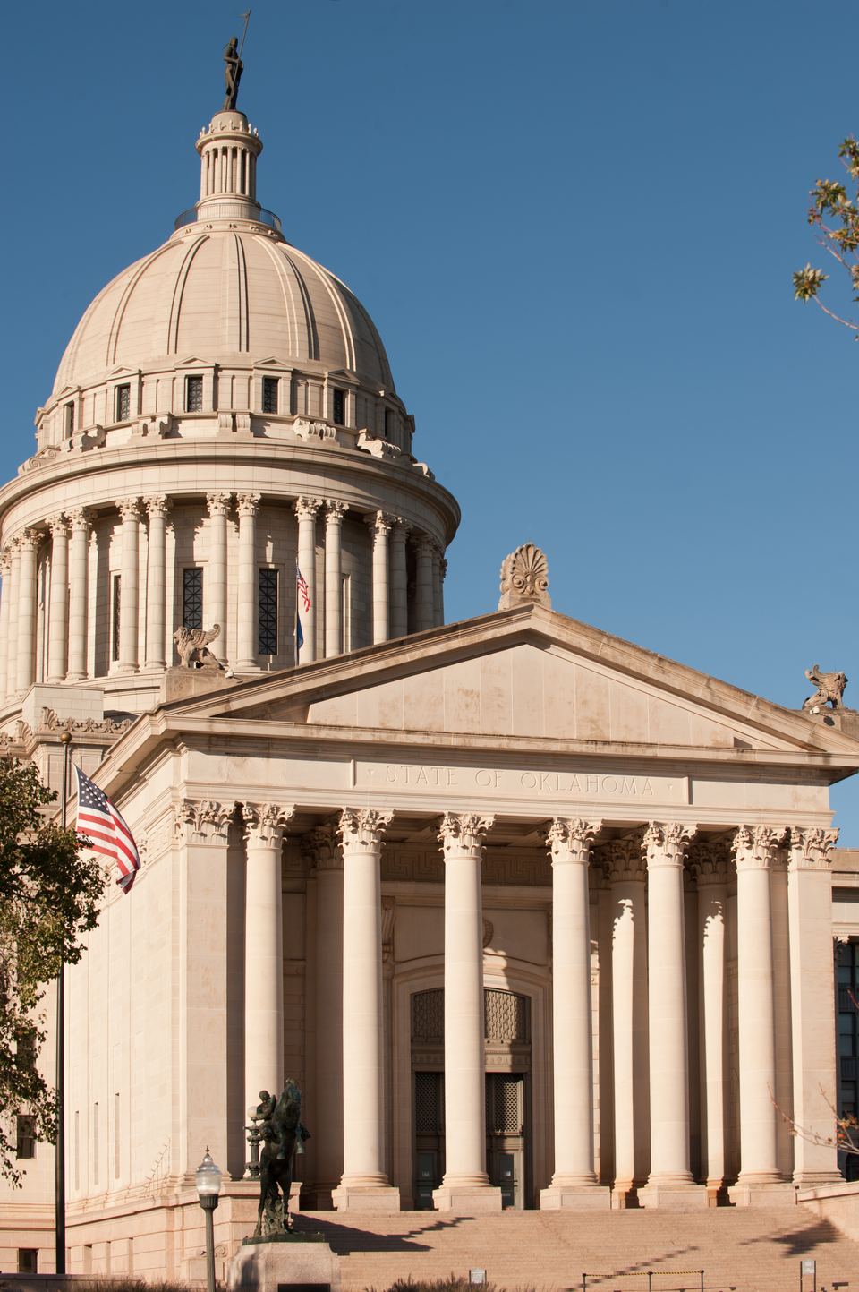 Columned south portico and dome of the Oklahoma State Capitol under a clear blue sky, with the U.S. flag and a bronze statue at the steps