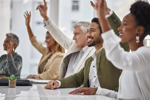 Diverse group of smiling people around a white table raising their hands to volunteer.